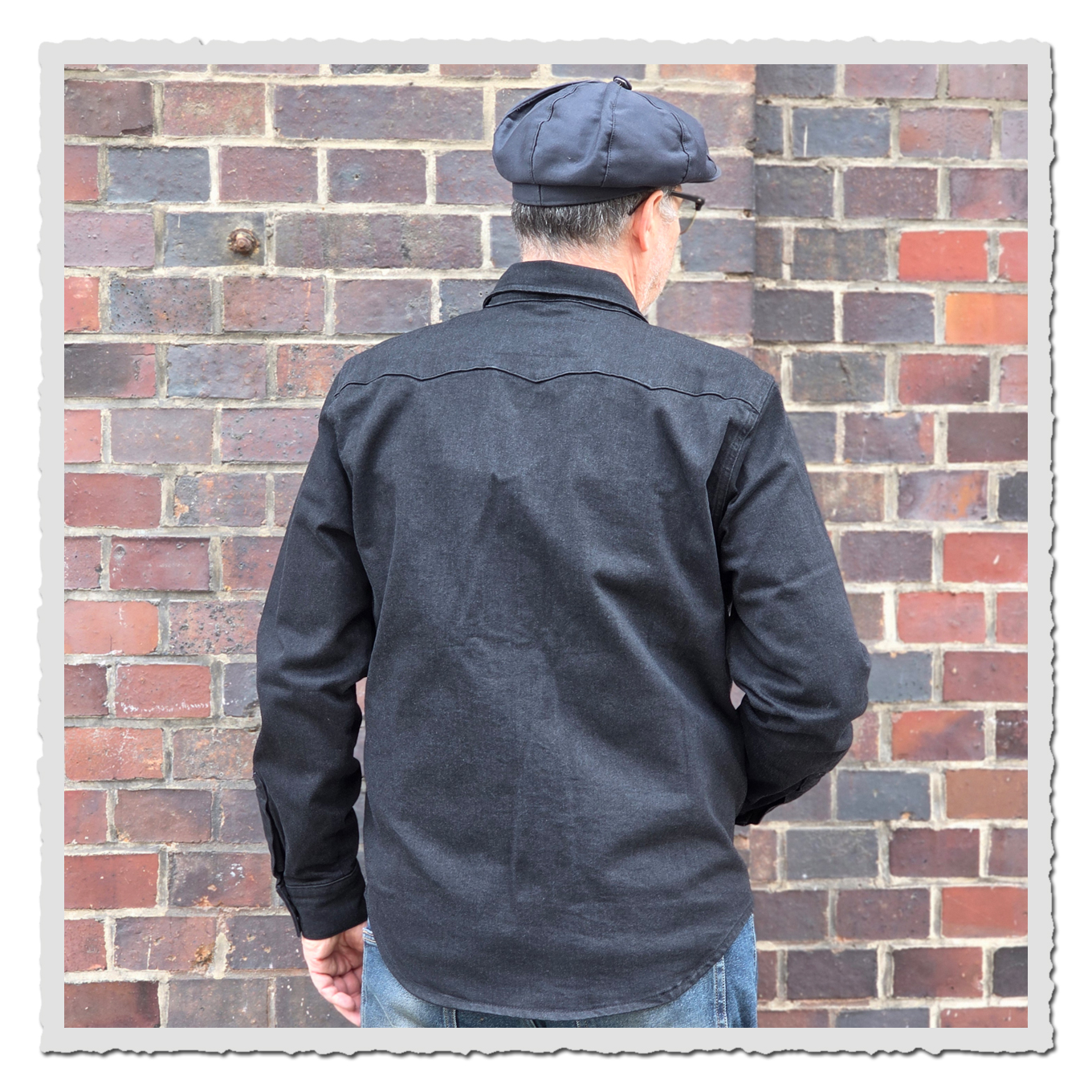 Person von hinten in dem schwarzem Hemd und dunkler Mütze vor roter Backsteinwand, leicht seitlich gedreht.